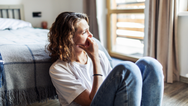 sad woman sitting on the floor in front of a bed looking out the window
