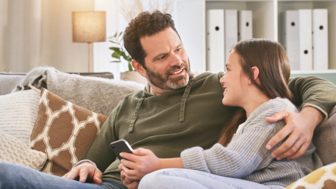 dad sitting on couch with his arm wrapped around his tween daughter as they talk
