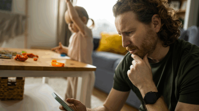 worried dad sitting and looking at his phone while little girl plays in background