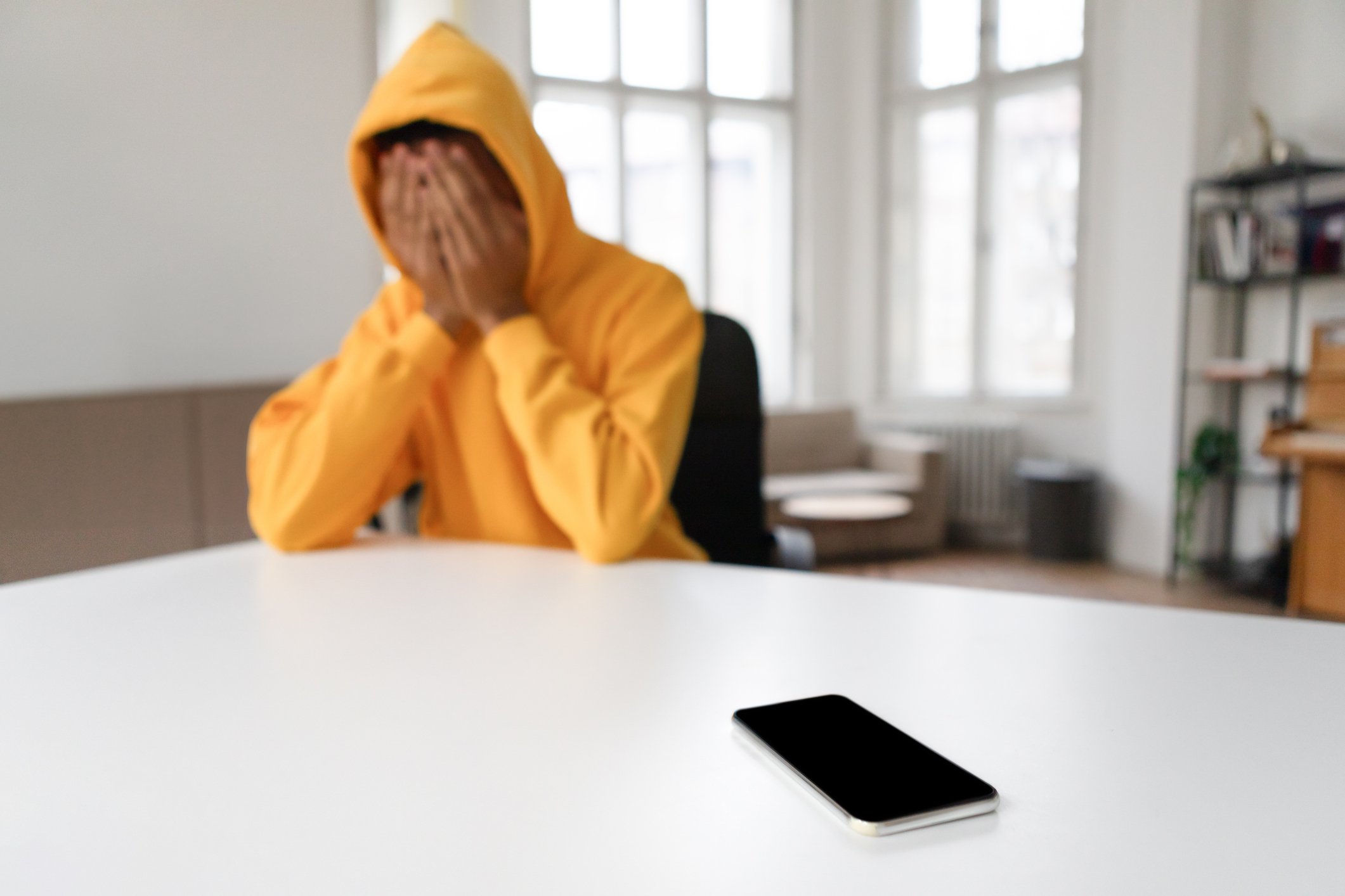 A teen boy in distress sits with their head in hands, a smartphone lying on a table