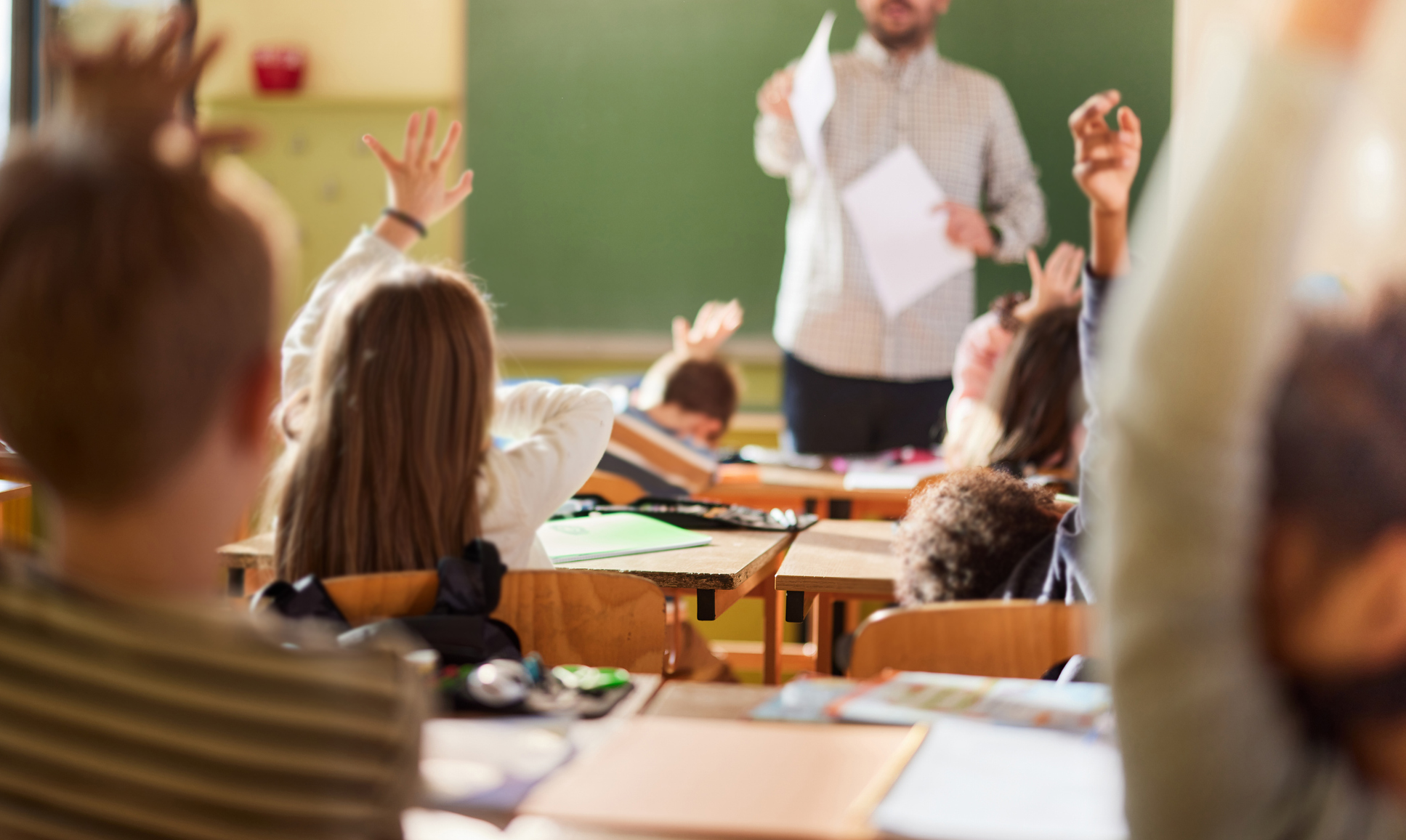Rear view of large group of school kids raising their hands to answer the teacher's question