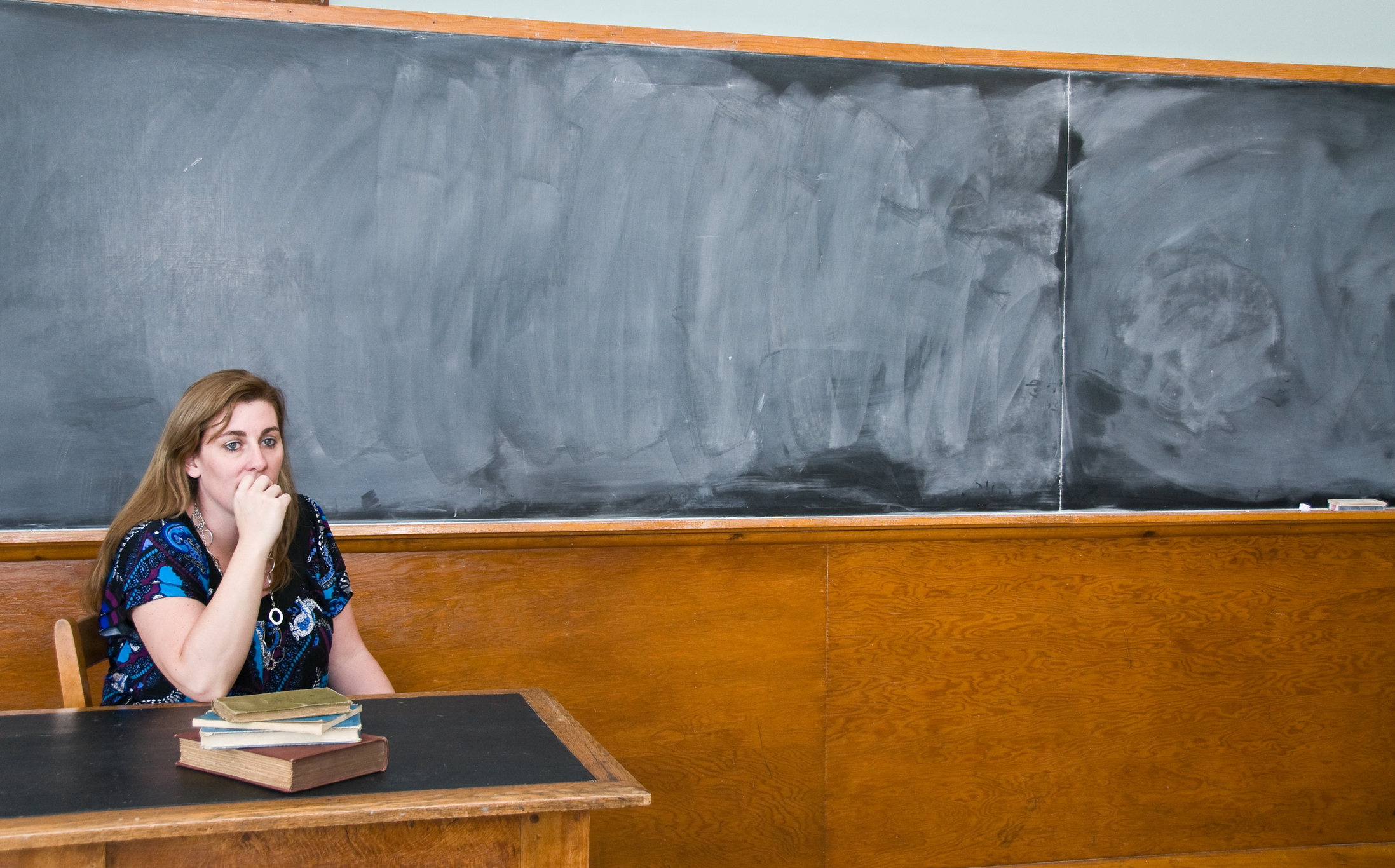 A teacher sitting at her desk looking worried with an old style blackboard behind her.