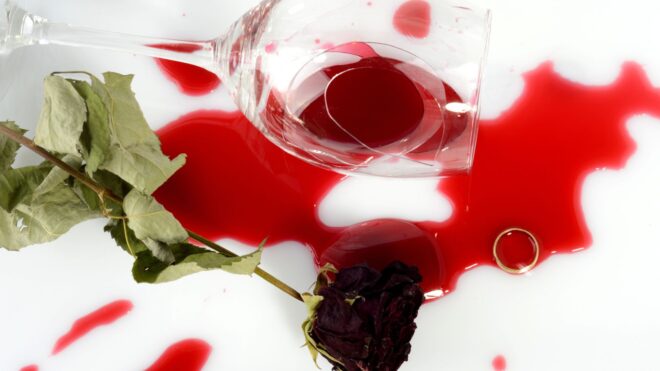 A glass tumbler of spilled red wine with a rose beside it and a wedding band