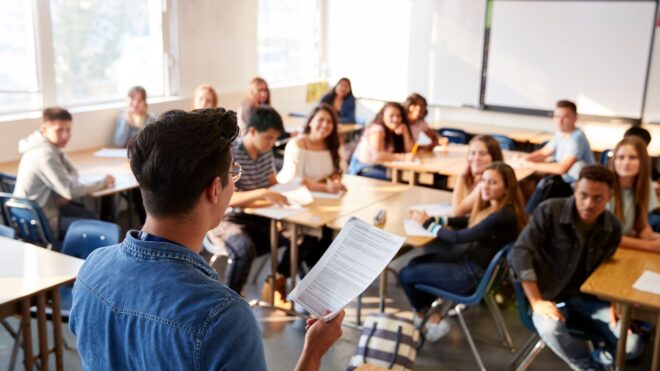 A male teacher standing up in front of their high school class full of students at desks