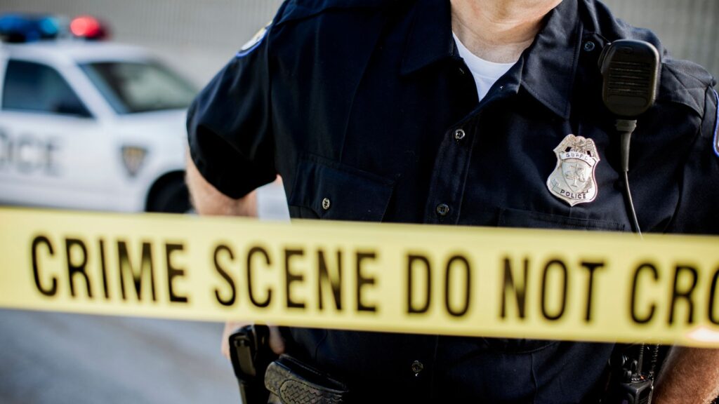 A police officer in uniform with a badge standing behind yellow crime scene tape with a police car parked behind him
