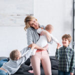 exhausted mother talking by smartphone and holding infant child while naughty siblings playing in room