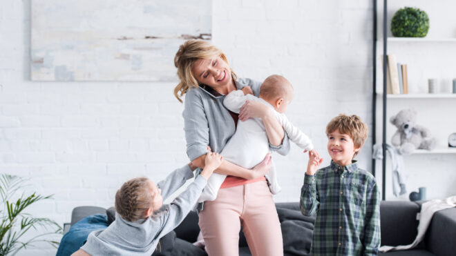 exhausted mother talking by smartphone and holding infant child while naughty siblings playing in room