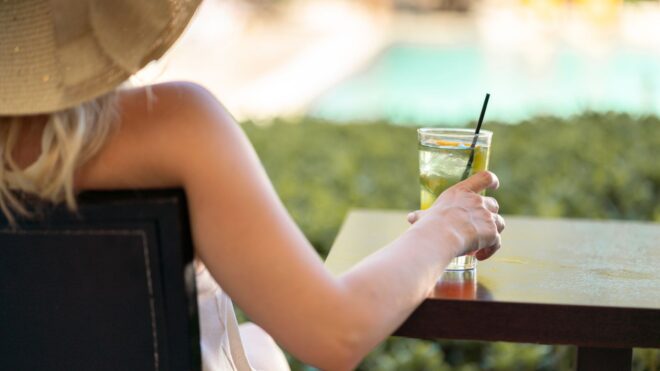 A close up shot of a woman in a beach hat sitting at a table with a drink looking out at a pool
