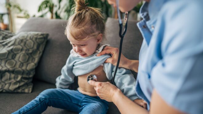 A healthcare worker examining a crying toddler with a stethoscope at home
