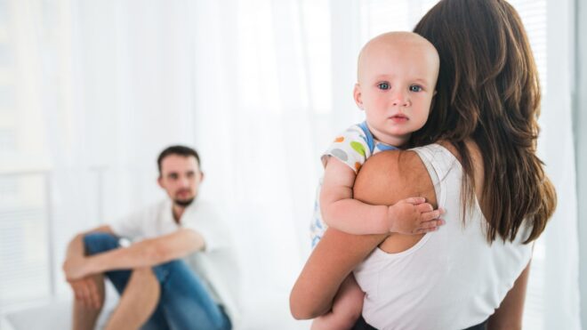 A mom holding her baby facing a man sitting down in the background
