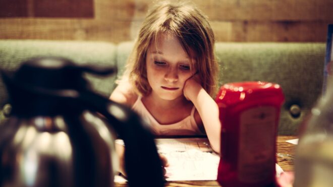 A photo of a young girl sitting alone at a restaurant looking sad with a ketchup bottle on the table