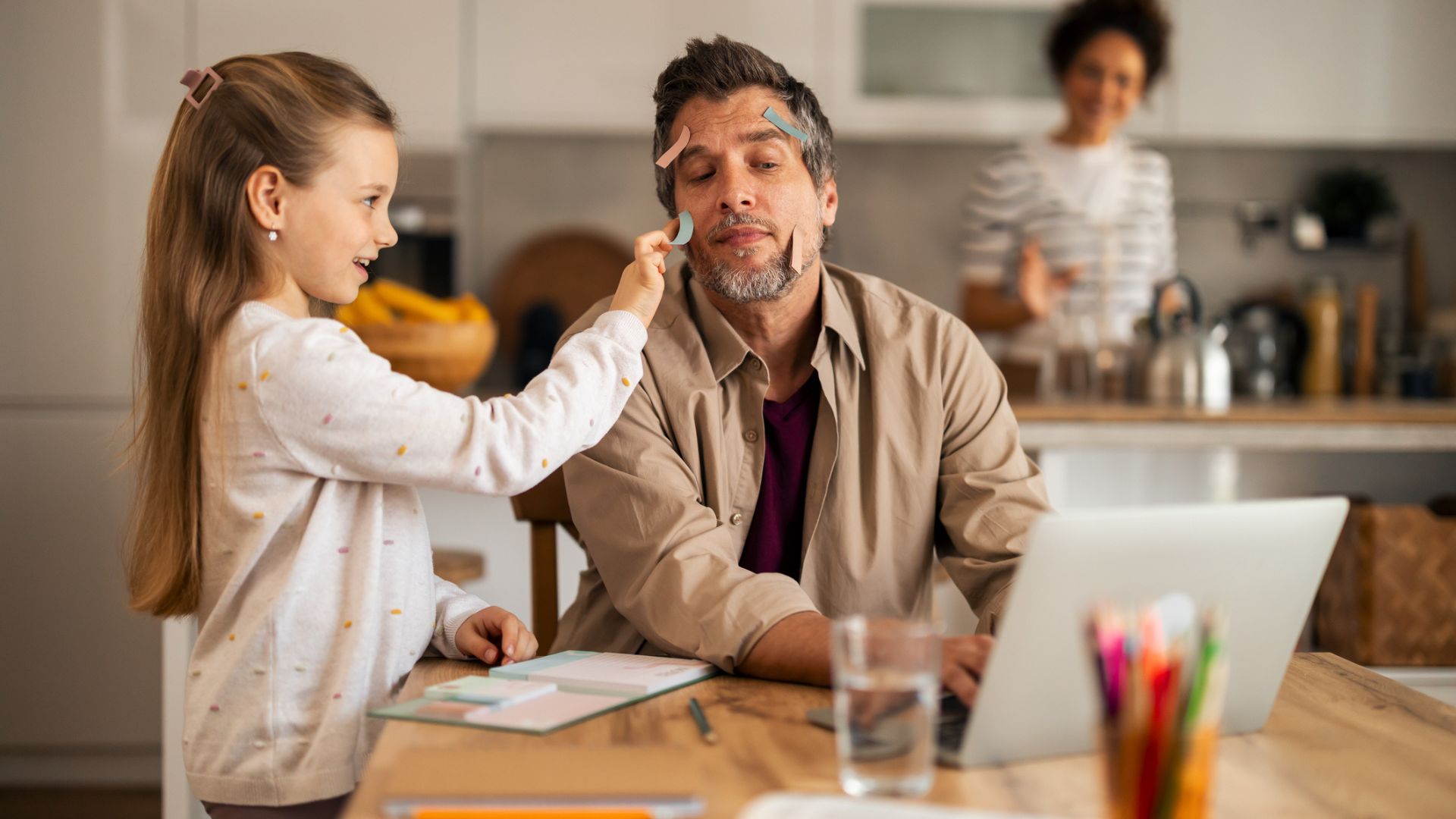 A dad sitting at the kitchen table on a laptop with his wife cooking behind him and a child being playful putting sticky notes on his face