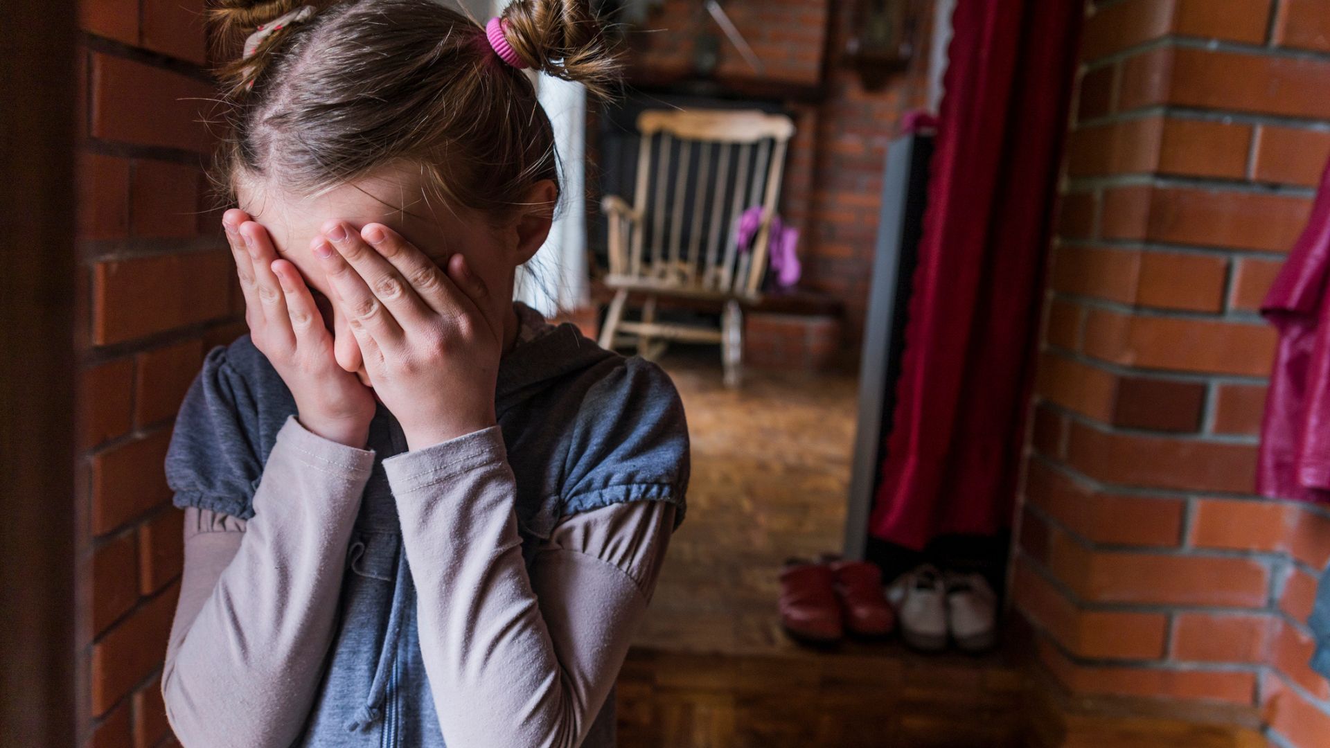 A young girl with pigtail buns covering her face scared outside of a brick room with a wood chair