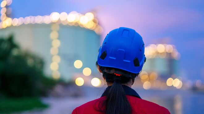 Woman in a blue safety helmet and red jacket overlooking a sight with lights and trees