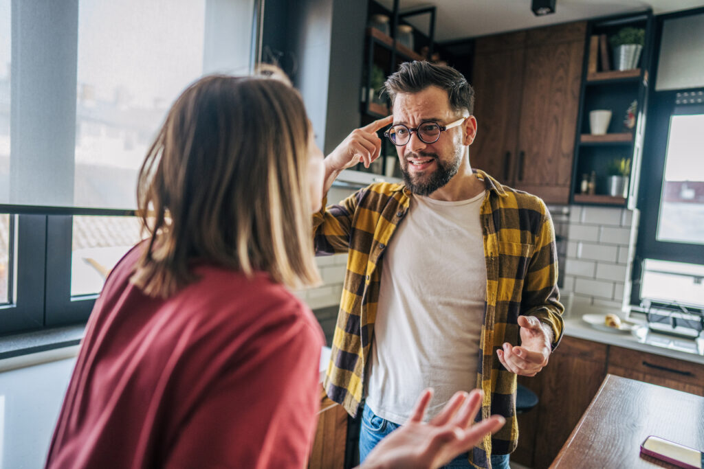 Boy and girl argument in the kitchen. He thinks that she is out of her mind, that she is crazy. He is not sure what is wrong. He puts his finger on his head.