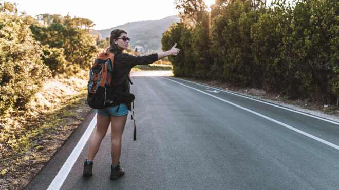 Female hiker wearing sunglasses and shorts, carrying backpack stopping by the side of country road, hitchhiking in the countryside