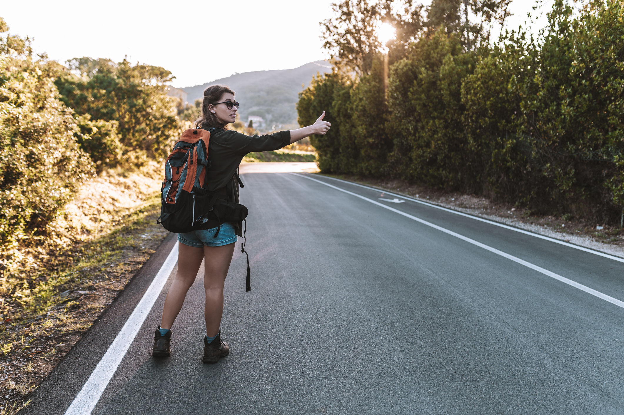 Female hiker wearing sunglasses and shorts, carrying backpack stopping by the side of country road, hitchhiking in the countryside