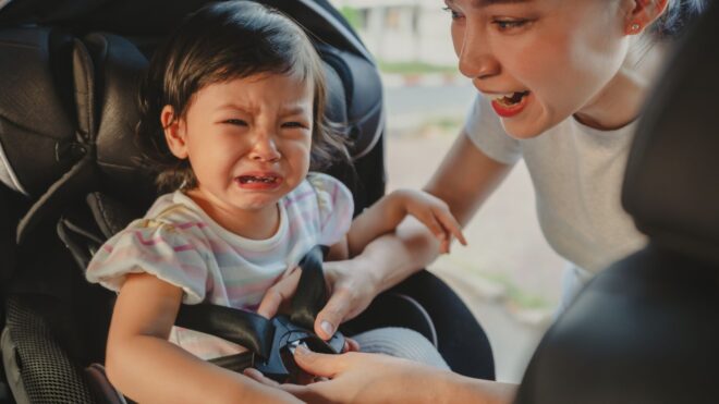 A mom buckling in a crying toddler girl into her car seat
