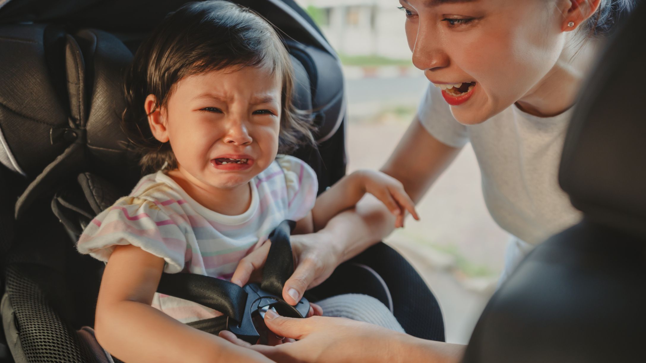 A mom buckling in a crying toddler girl into her car seat