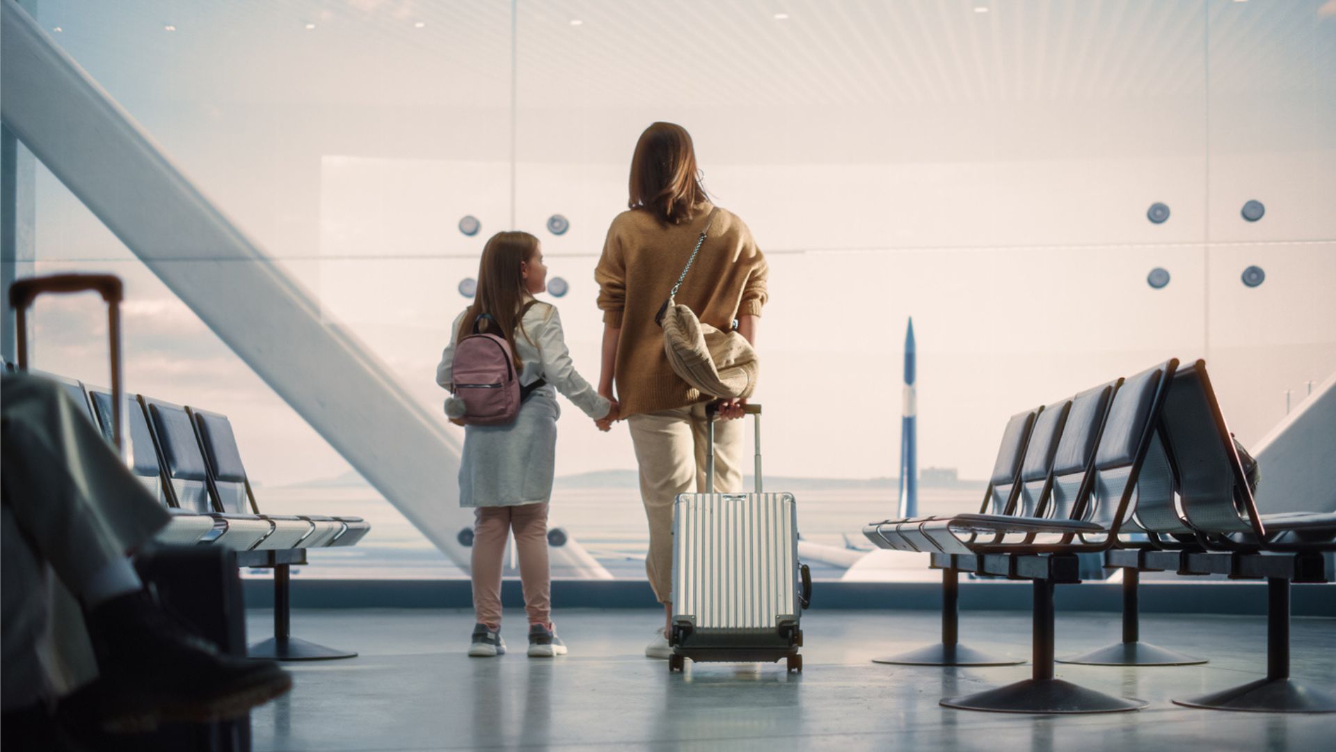 A mother and daughter hold hands in an airport terminal while holding a suitcase behind them