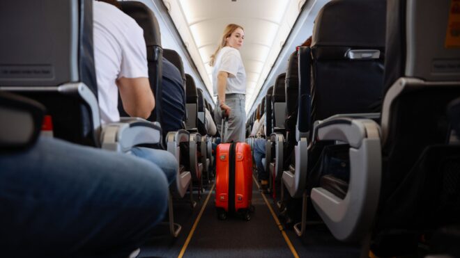 A woman in the aisle of an airplane with an orange rolling suitcase