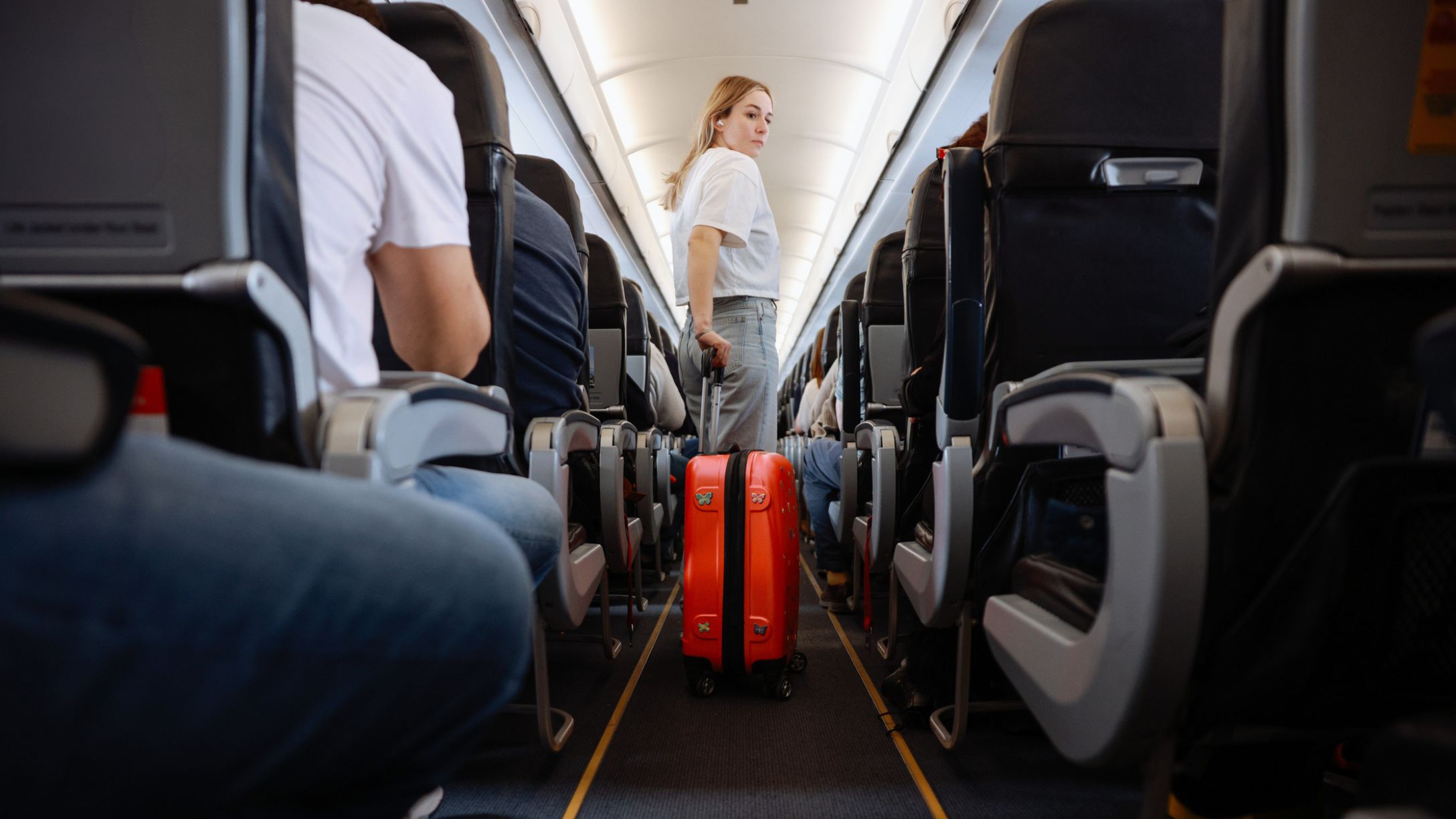 A woman in the aisle of an airplane with an orange rolling suitcase