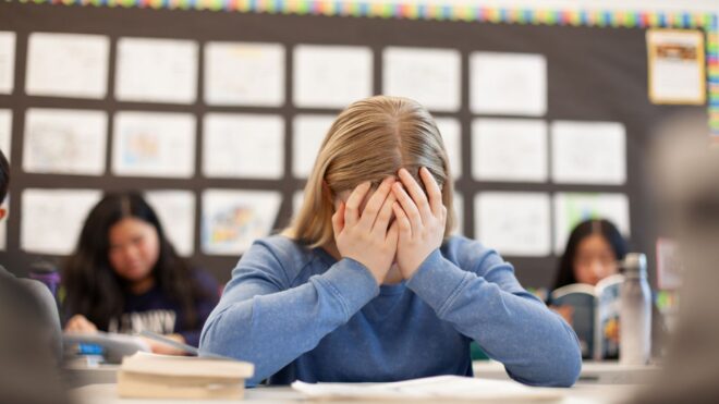 An elementary school student sitting at her desk in class with her hands covering her face
