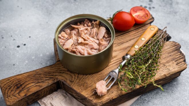 An opened can of tuna on a rustic wooden cutting board with herbs, a knife, and tomatoes