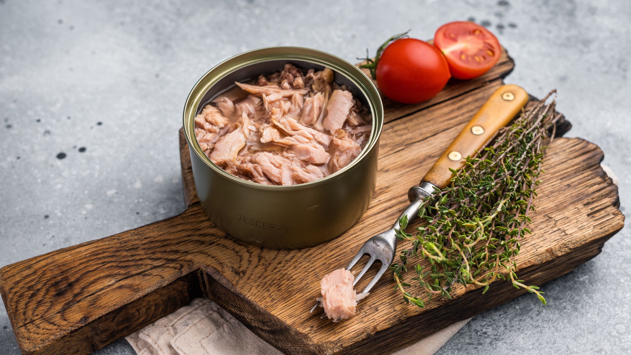 An opened can of tuna on a rustic wooden cutting board with herbs, a knife, and tomatoes