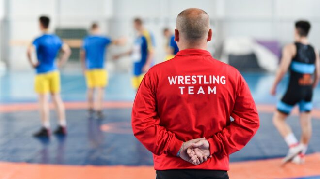 Coach in a red jacket observing wrestlers during practice