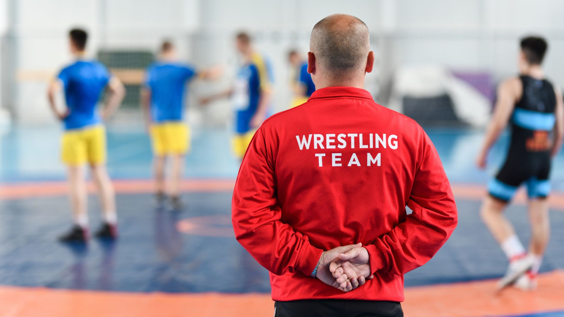 Coach in a red jacket observing wrestlers during practice