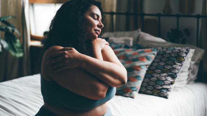 Curly haired overweight young woman in blue top and shorts with satisfaction on face accepts curvy body shape in stylish bedroom.