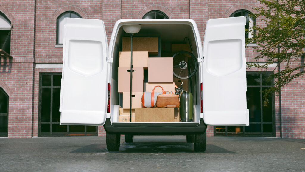 White moving van parked in front of building. The back doors to the van are open showcasing lots of cardboard boxes, bags and some furniture. 