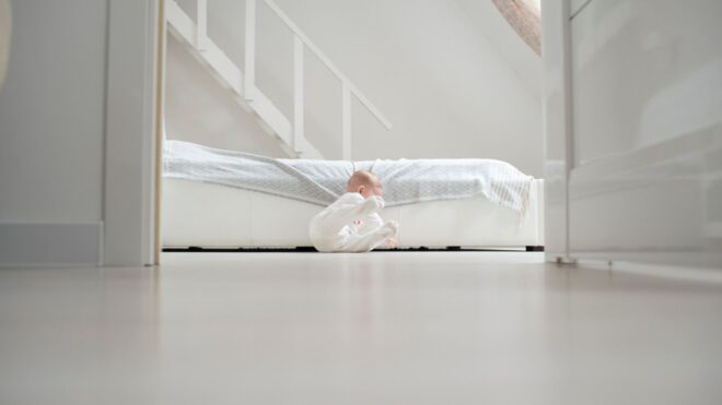 A baby falls off a bed on the floor in a white bedroom