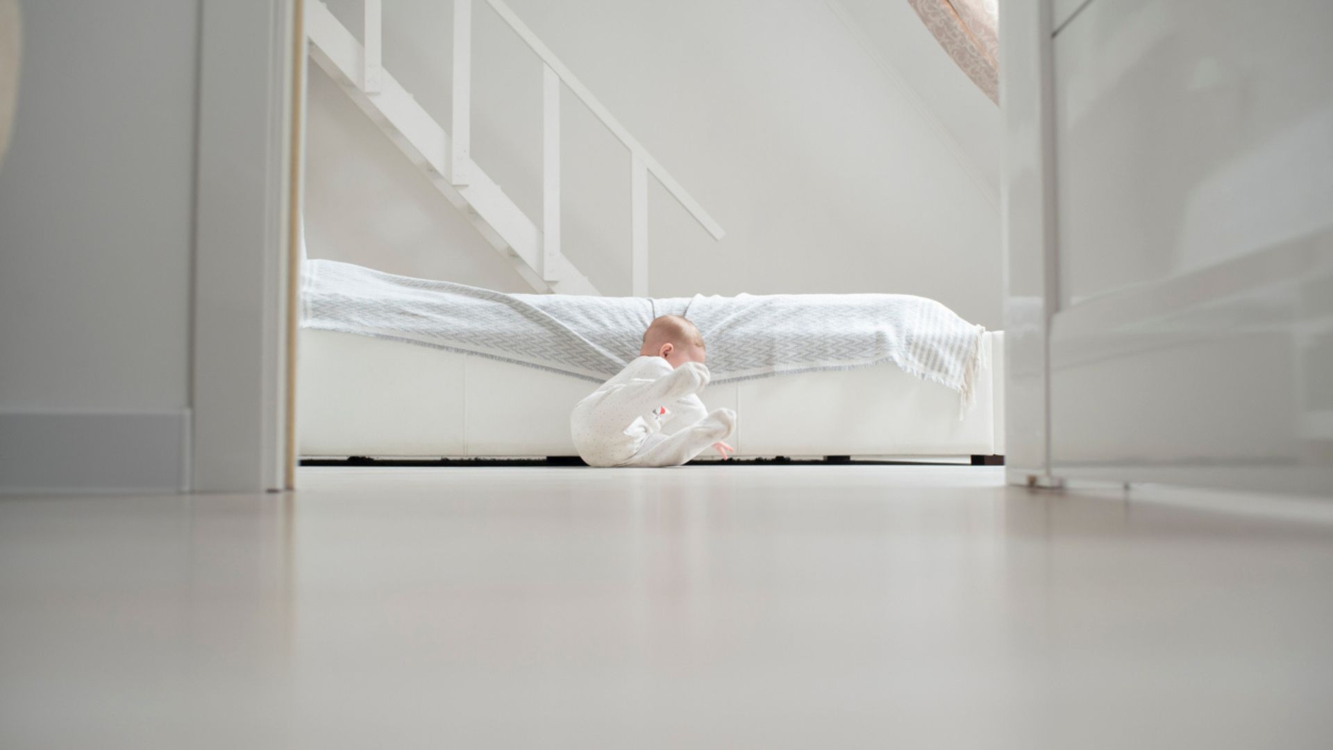 A baby falls off a bed on the floor in a white bedroom