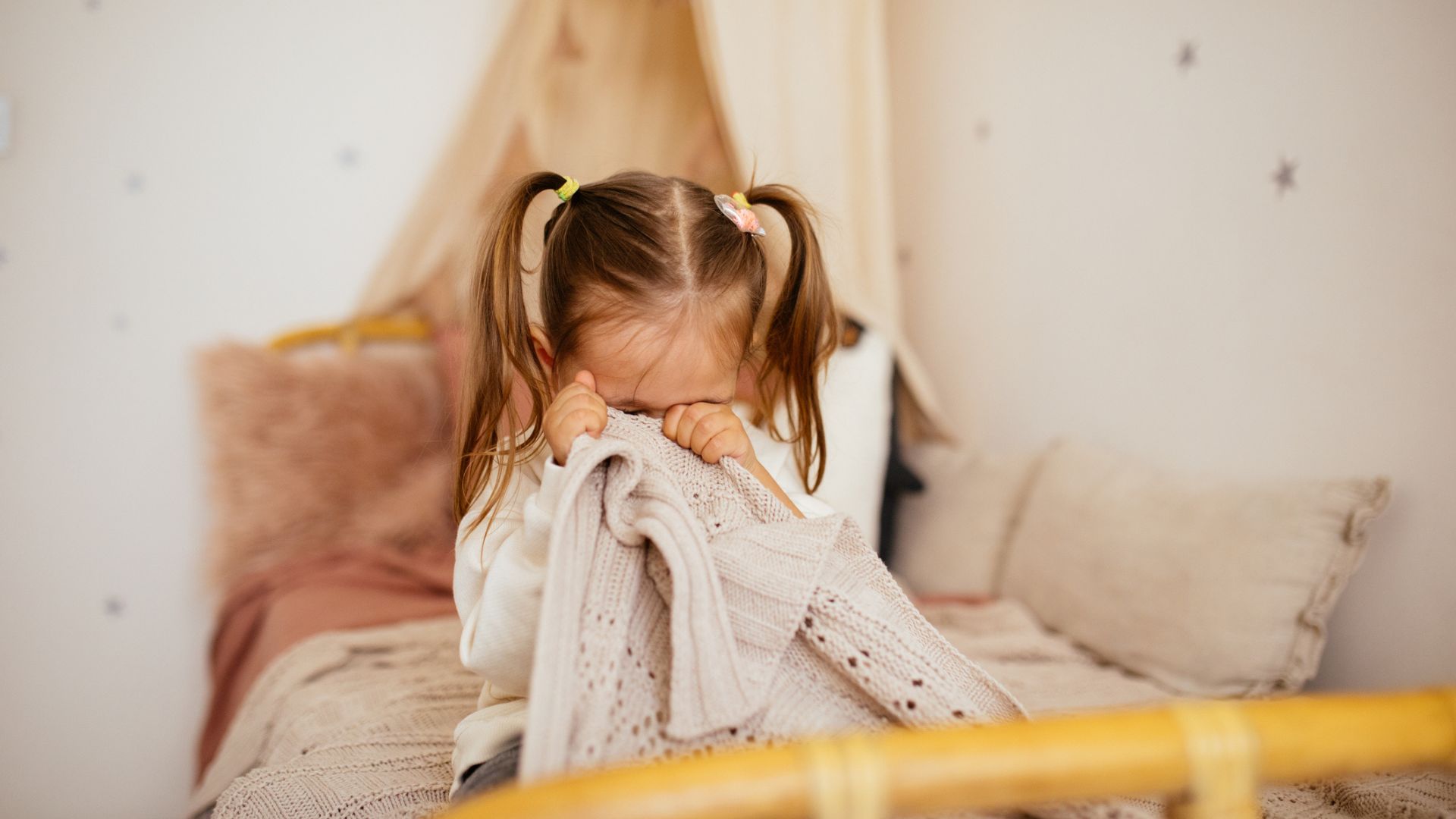 A crying little girl with pigtails in bed covering her face with a blanket