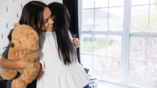 A grown woman holding a large teddy bear and hugging a teen girl