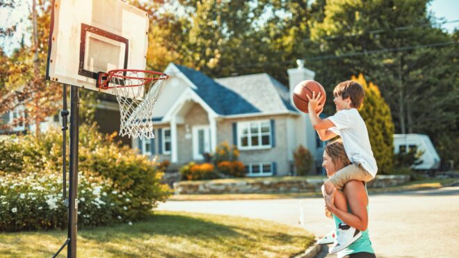 A photo of a kid playing basketball on an older teen's shoulder in a neighborhood in front of a house