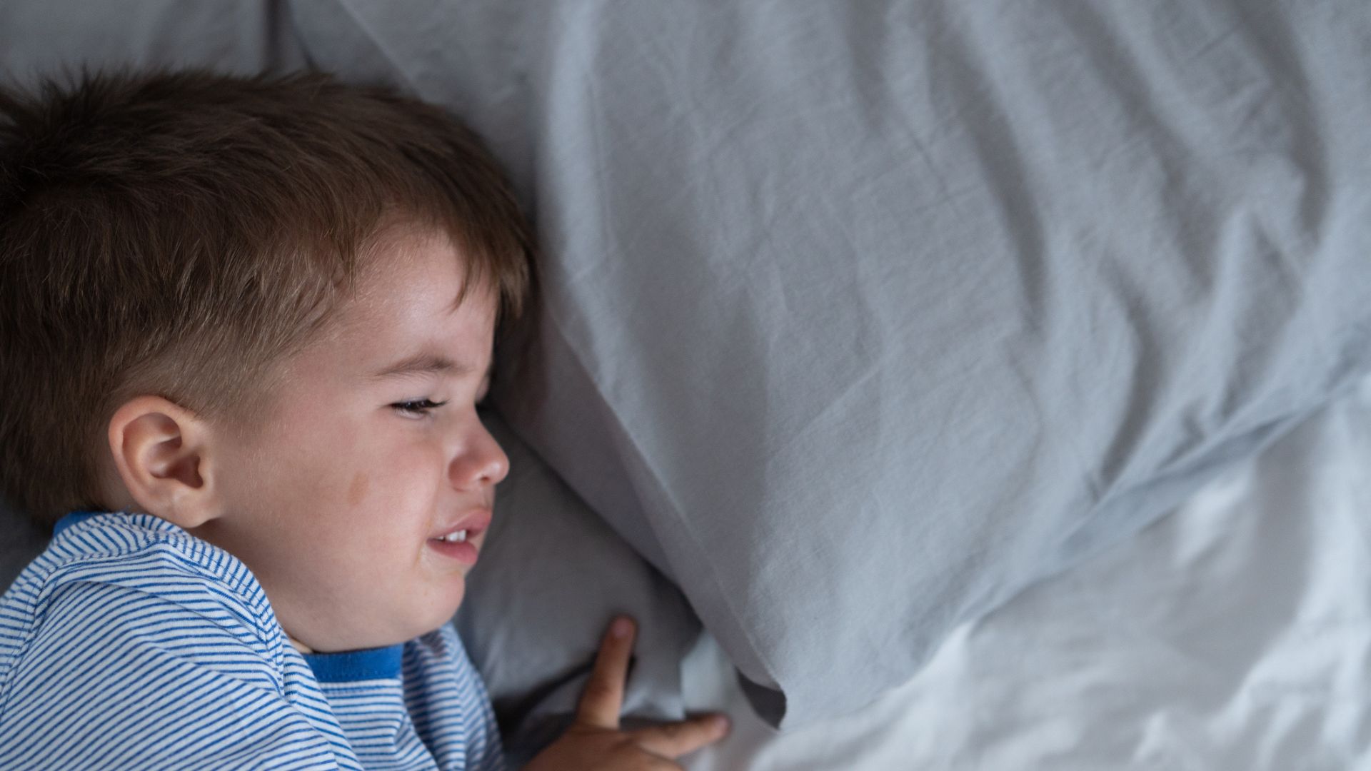 A toddler in a blue striped shirt cries in bed