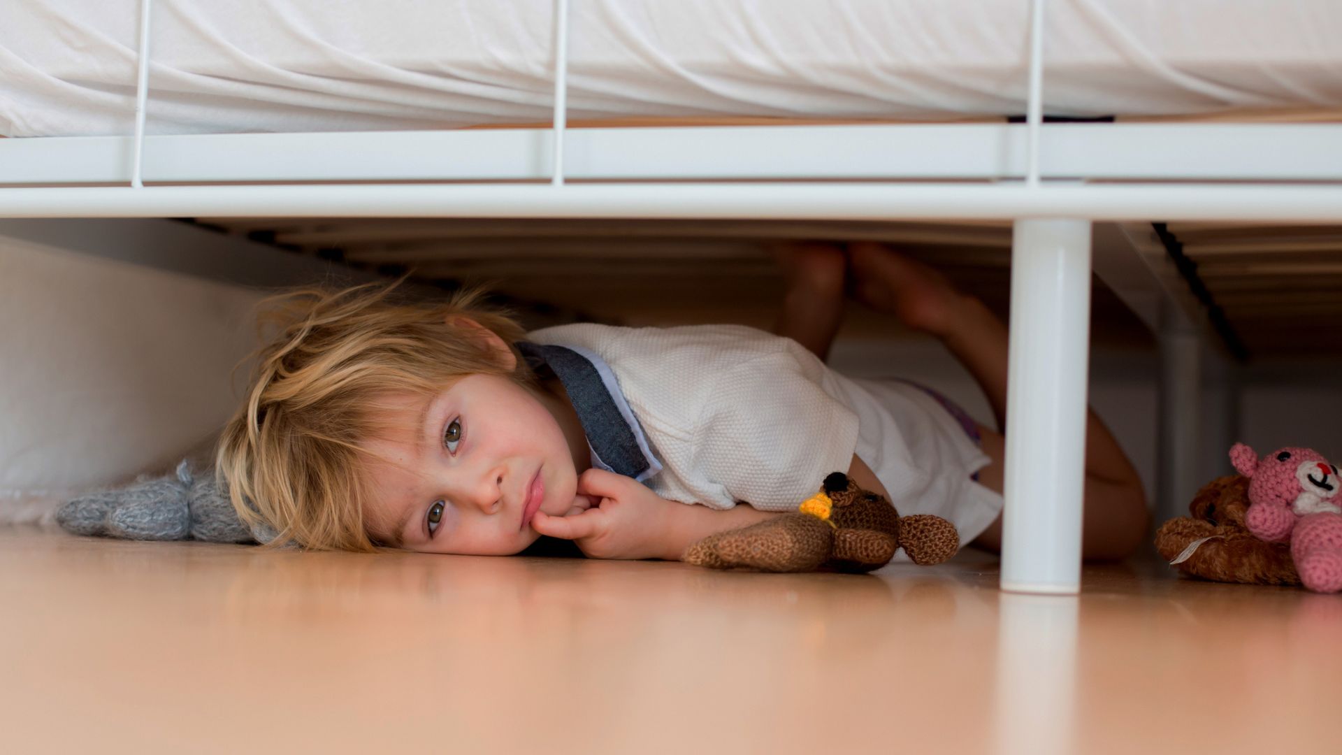 A young boy laying underneath his bed with some toys nearby
