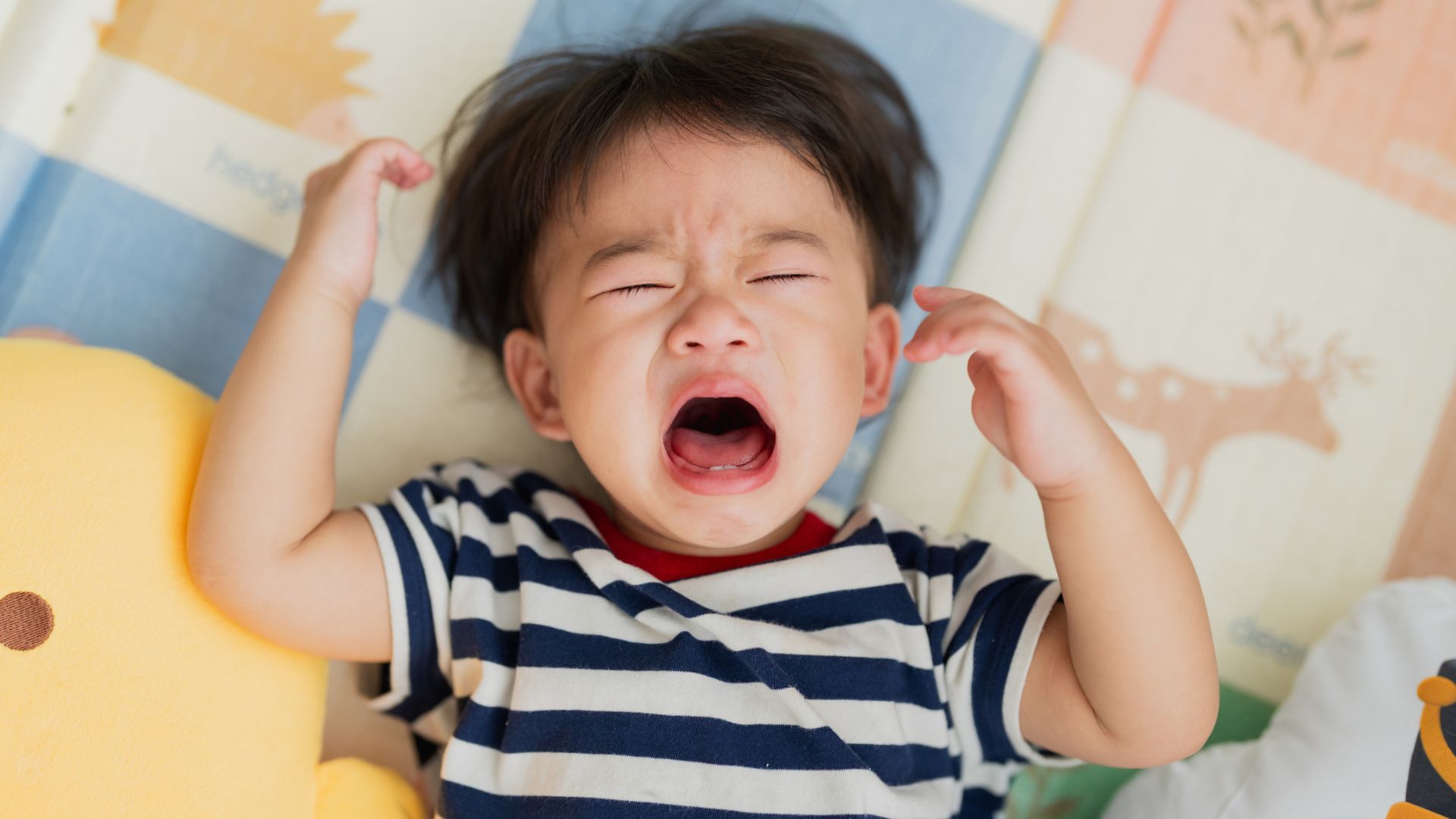 An Asian toddler crying with their eyes closed on a colorful play mat