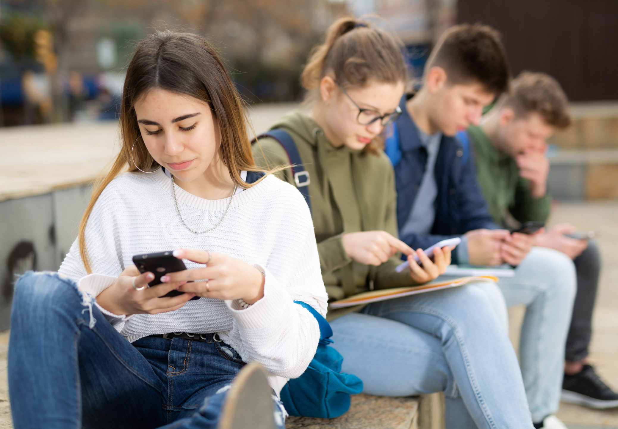 Portrait of girl teenager addicted in phone sitting with friends on bench outdoors