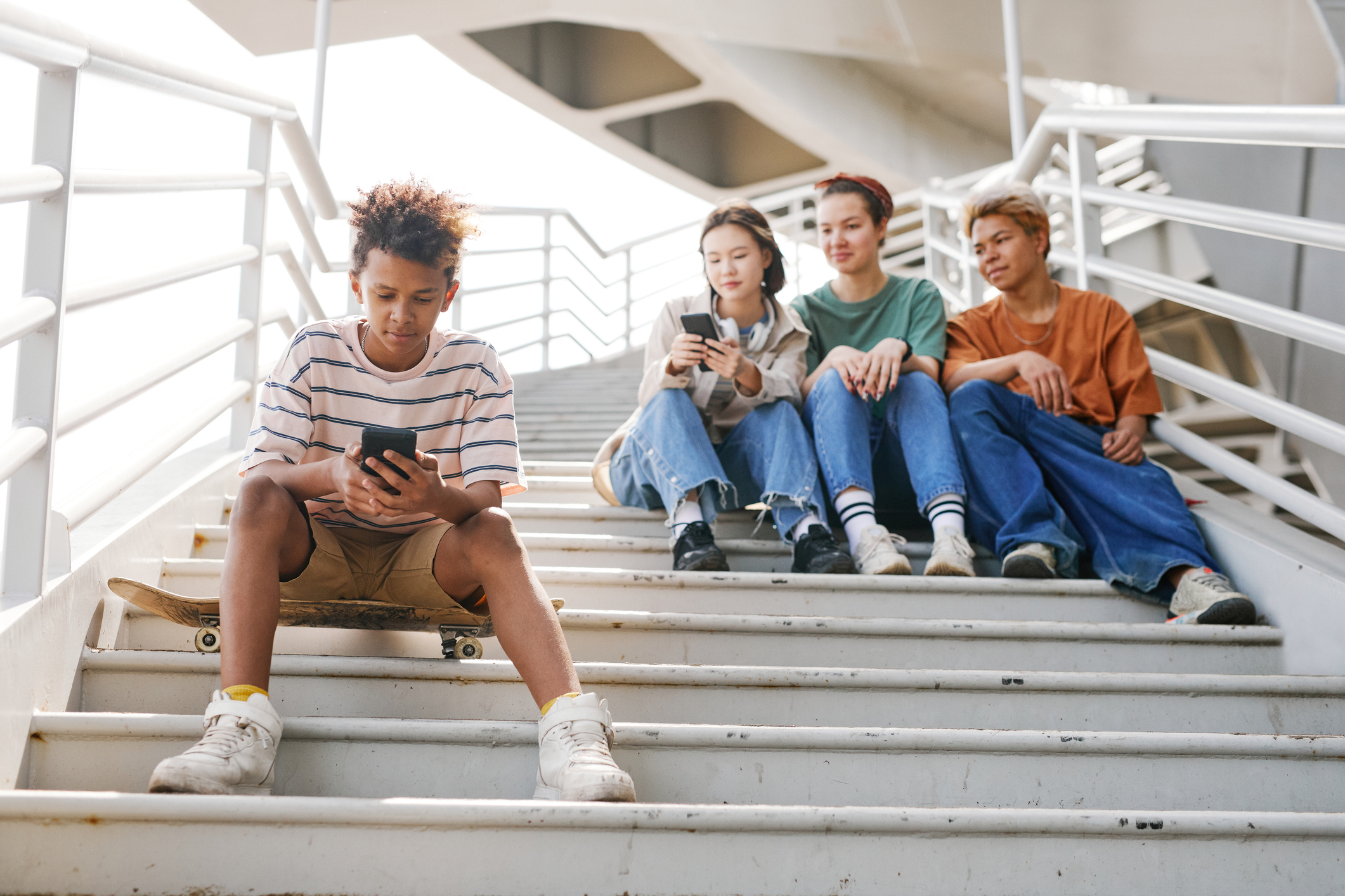 Full length portrait of teenage boy using smartphone outdoors while sitting on metal stairs with group of friends in background, copy space