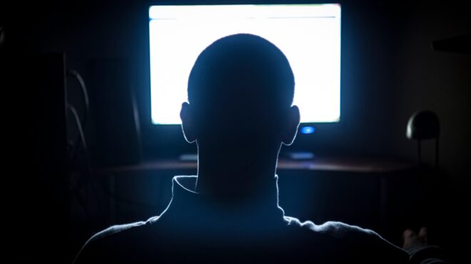 A man sits in a dark room in front of a bright screen