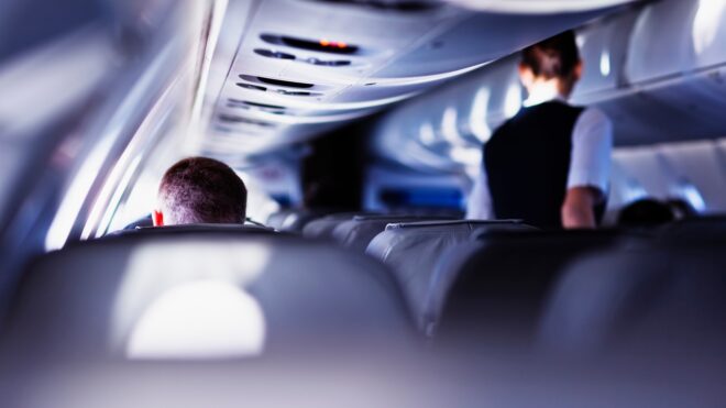 Flight attendant walks down the main aisle of a plane towards a man.