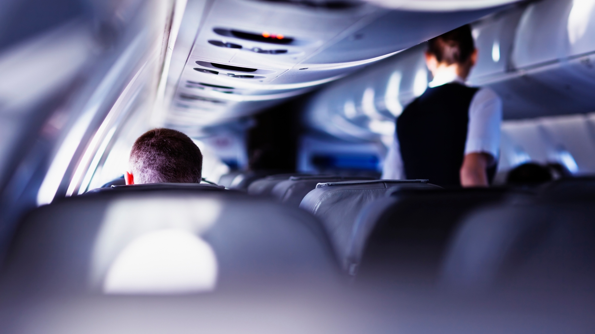 Flight attendant walks down the main aisle of a plane towards a man.