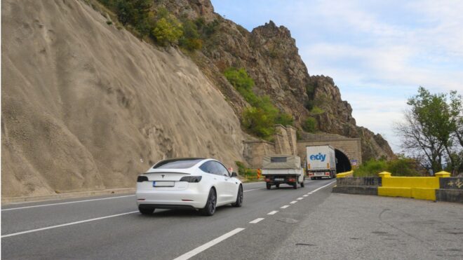 A white Tesla on a highway next to a rocky cliff with a few cars ahead of them