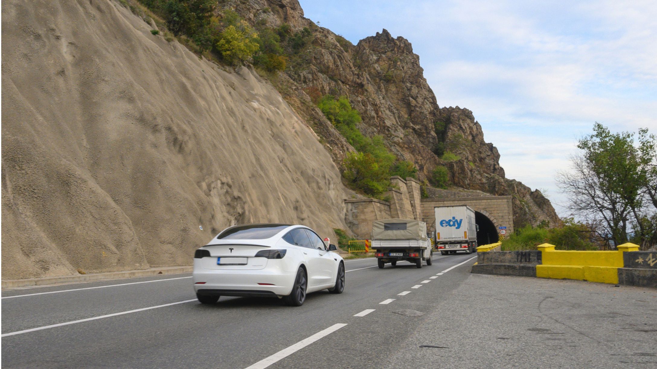 A white Tesla on a highway next to a rocky cliff with a few cars ahead of them