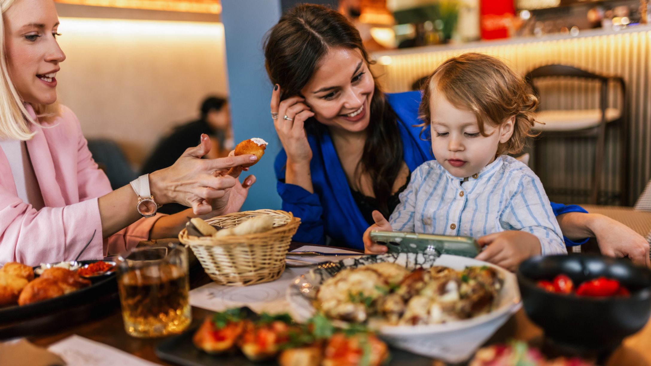 Two women gathered at a restaurant with a table full of a food and a child on an tablet