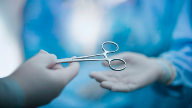 Hand of nurse passing medical tool to doctor palm of hand wearing protective workwear in medical surgery operation room in hospital with patient incidental people background stock photo
